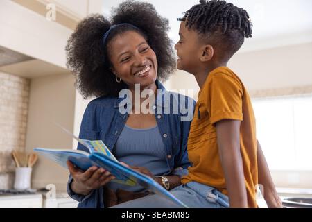 Mère afro-américaine et son fils lisant un livre d'images coloré sur le comptoir de cuisine souriant Banque D'Images