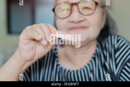 Heureuse dame senior tient et montre son nouvel implant dentaire et sa mâchoire avec une paire de dents. Concepts des problèmes de santé bucco-dentaire des seniors et soins dentaires Banque D'Images