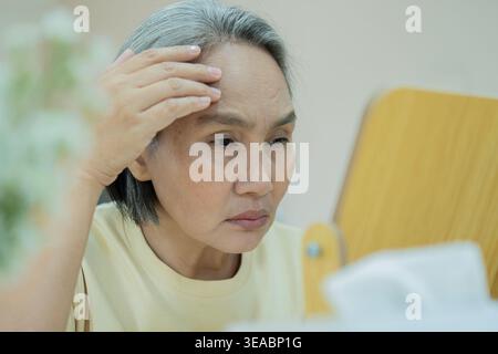 Asie les personnes adultes d'âge moyen la femme âgée regarde le miroir peau sèche visage toucher la main sur le front joue stress triste à la maison. Soins de santé aînés la beauté vieillissante est Banque D'Images