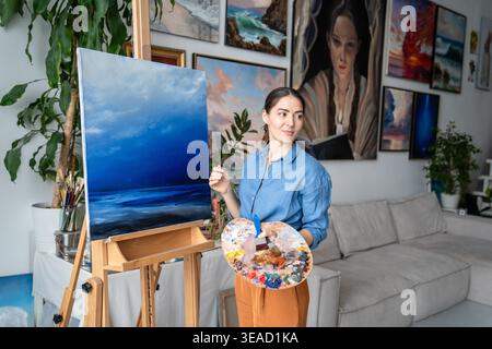 Artiste femme créative poursuivant un passe-temps d'art, peintre féminin positif réfléchi dessinant image sur toile avec palette et pinceau au studio loft, créateur professionnel appréciant l'art thérapie. Banque D'Images