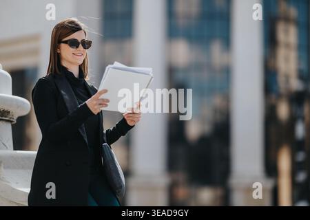 Femme d'affaires lisant des documents à l'extérieur en ville tout en portant des lunettes de soleil et une tenue noire intelligente Banque D'Images