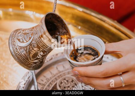 Mains de femme versant du café turc chaud frais d'un cezve en métal classique dans une petite tasse traditionnelle, montrant une boisson culturelle Banque D'Images