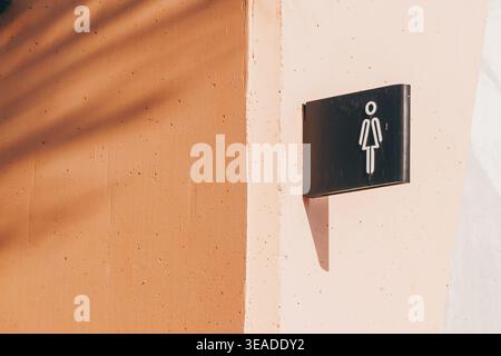Enseigne des toilettes pour femmes attachée à un mur de couleur pêche, représentant un symbole féminin minimaliste et des ombres et lumières contrastées Banque D'Images