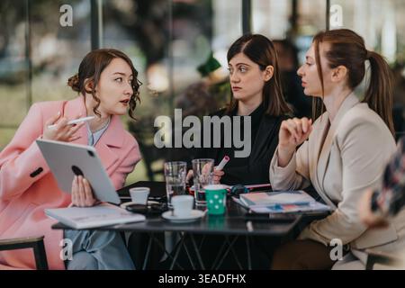 Trois femmes d'affaires ayant une discussion autour de documents et de tablettes lors d'une réunion de café Banque D'Images