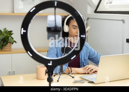 Personne non binaire enregistrant un podcast à son bureau à l'aide d'un éclairage annulaire LED, d'un micro et d'un casque Banque D'Images