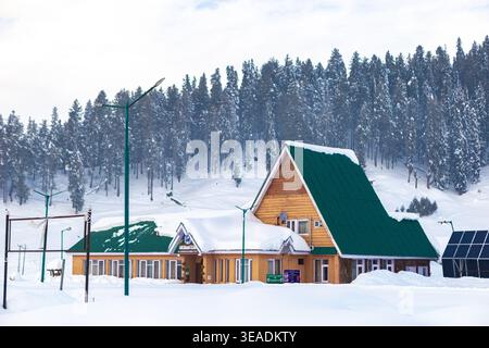 Vue aérienne d'un bâtiment en bois avec un toit vert niché dans un paysage enneigé, encadré par des pins denses, créant une scène hivernale sereine, Gulmarg, Banque D'Images