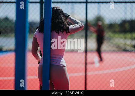 Femme en tenue athlétique appuyée sur un poteau à un terrain de sport extérieur rouge derrière une clôture à maillons de chaîne Banque D'Images