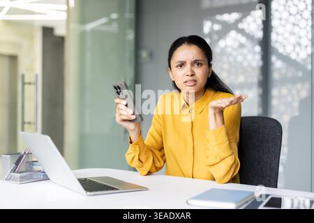 Femme au bureau tenant un smartphone, haussant les épaules dans la frustration et la confusion sur un problème de technologie ou de connexion, regardant vers la caméra avec une expression ennuyée et perplexe Banque D'Images