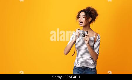 Jeune femme tient joyeusement un appareil photo devant un fond jaune vibrant Banque D'Images