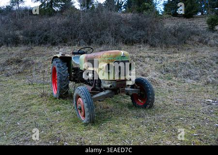 Vieux tracteur rouillé au ralenti dans un champ rural herbeux avec des arbustes et des arbres à l'arrière-plan, probablement des terres agricoles abandonnées ou inutilisées. Banque D'Images