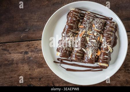 Gros plan photo de délicieuses crêpes à la sauce au chocolat et saupoudrées d'amandes sur une plaque blanche sur une planche de bois rustique. Crêpes de dessert fines, rouleau Banque D'Images