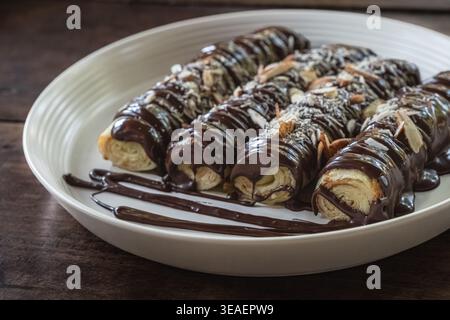 Gros plan photo de délicieuses crêpes à la sauce au chocolat et saupoudrées d'amandes sur une plaque blanche sur une planche de bois rustique. Crêpes de dessert fines, rouleau Banque D'Images