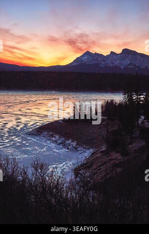 Coucher de soleil sur le lac gelé d'hiver avec montagnes enneigées et pins Banque D'Images
