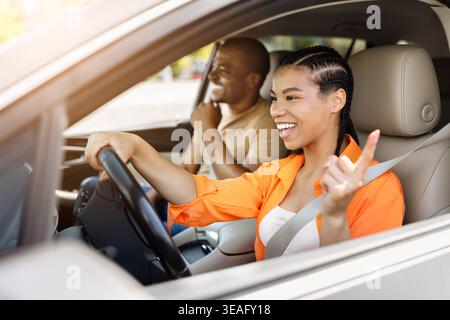 Heureuse jeune femme noire conduisant la voiture et faisant des gestes avec le doigt tout en souriant Banque D'Images