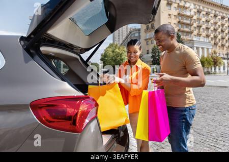 Heureux jeune couple noir chargeant des sacs à provisions colorés dans le coffre de la voiture Banque D'Images