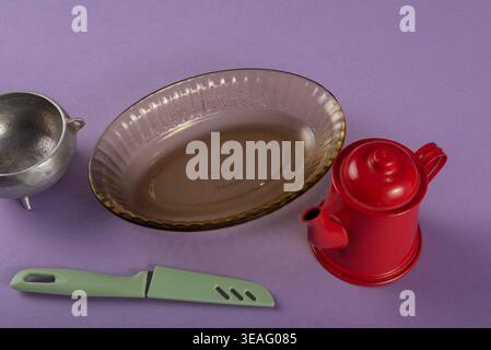 Nature morte avec une théière rouge, un bol argenté, un couteau vert clair et un plat de cuisson en verre. L'éclairage doux du studio met en valeur les textures. Tiré sur une lavande Banque D'Images