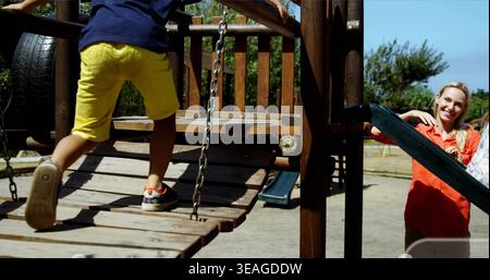 Garçon traversant en short jaune chemise marine sur pont de parc en bois, femme en chemisier orange regardant Banque D'Images
