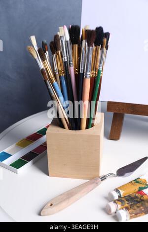 Pinceaux, couteau, peintures et chevalet avec toile sur table près du mur bleu, gros plan Banque D'Images