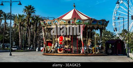 Un carrousel de style vintage avec des panneaux peints se trouve dans un parc près de palmiers et d'une grande roue à Olbia, en Sardaigne, sous un ciel bleu clair. Banque D'Images