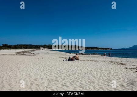 Les gens se détendent sur le sable blanc de la plage de Bados à Olbia, en Sardaigne, bordée par les eaux méditerranéennes bleues et la végétation côtière verte sous un SK clair Banque D'Images