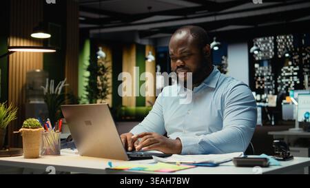 Homme afro-américain fait la pensée critique et la relecture avec des papiers avant de transcrire des données sur ordinateur portable, assis dans le bureau la nuit. Travailleur documentant les idées importantes. Banque D'Images