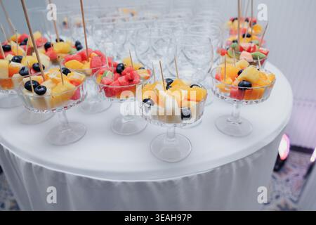 Tasses de fruits individuelles dans des bols en cristal sur la table ronde sur pied, brochettes délicates avec melon et baies, présentation raffinée Canap pour cocktail Banque D'Images