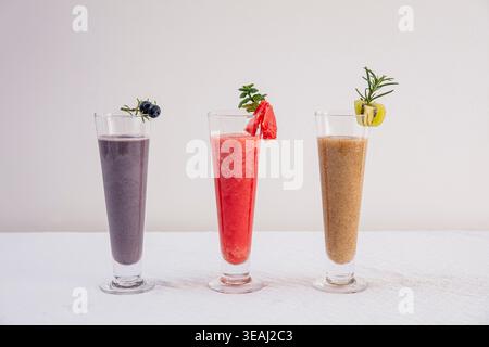 Trois grands verres de smoothies différents, chacun garni d'une garniture unique, reposent sur une nappe blanche dans un cadre lumineux et moderne. Banque D'Images