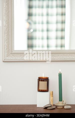 Une bougie allumée dans un bocal en verre se trouve sur une table en bois, entourée de cailloux et d'un bougeoir, devant un mur blanc et un miroir argenté. Banque D'Images