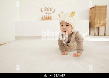 Un bébé dans un chapeau de mouton repose sur un sol blanc, regardant autour dans une pièce ensoleillée. Banque D'Images