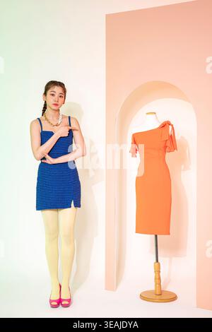 Une jeune femme dans une robe bleue se tient à côté d'une robe orange sur un mannequin, tous deux dans une pièce blanche neutre. Banque D'Images