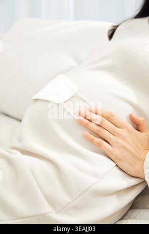 Une femme portant un masque facial est assise sur un canapé, berçant son ventre enceinte. Banque D'Images