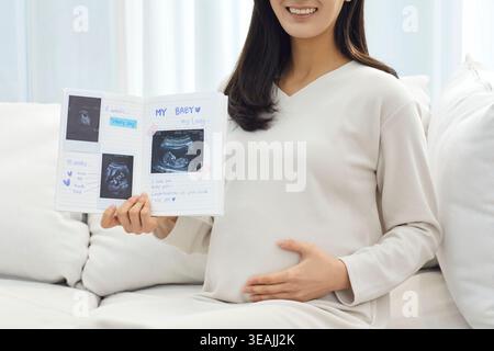 Une femme assise sur un canapé, tenant un livre d'échographie et souriant tout en montrant les photos échographiques de son bébé. Banque D'Images