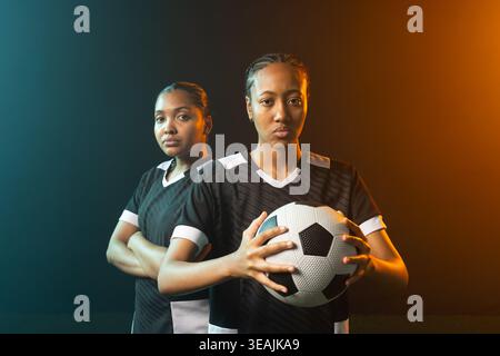 Joueuses afro-américaines posant dans un studio tenant un ballon de football dans un kit noir sous un éclairage gel Banque D'Images