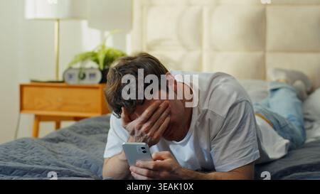 Homme caucasien inquiet allongé sur le lit à la maison tenant un téléphone portable saisissant la tête bouleversé homme choqué problème gars déçu par l'échec d'erreur de smartphone Banque D'Images