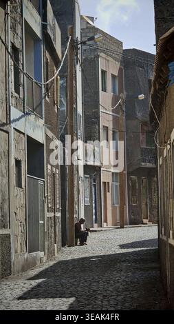 Une étroite ruelle vide avec des pavés et de grands bâtiments partiellement délabrés, un homme assis sur la marche devant une entrée de maison, Ribeira Grande Banque D'Images