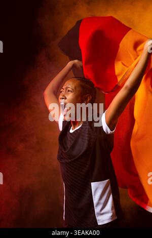 Femme afro-américaine criant, tenant le drapeau rouge-orange dans le studio en maillot de sport avec brume fumée. Solo, dramatique, dynamique, vibrant, célébration, spo Banque D'Images
