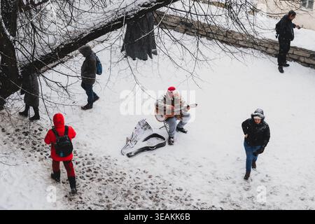 Tallinn, Estonie - 03 janvier 2025 : vue d'un guitariste solitaire faisant la sérénade des passants sur la place enneigée de l'hôtel de ville, une touche de manteaux rouges vibrants Banque D'Images