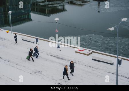 Tallinn, Estonie - 03 janvier 2025 : vue de personnages marchant le long du sentier enneigé à côté de l'eau gelée, reflétant la façade vitreuse du bâtiment Banque D'Images