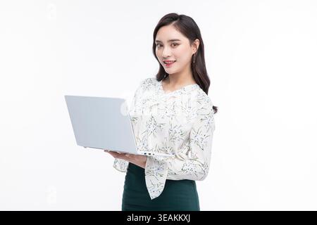 Une jeune femme dans une blouse blanche et une jupe sombre travaille sur un ordinateur portable dans une pièce lumineuse et neutre. Banque D'Images