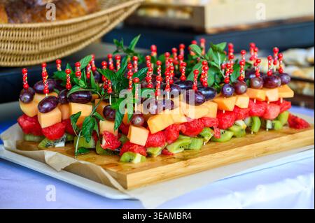 Rangées de brochettes de fruits frais colorées avec raisins, melon, pastèque et kiwi, garnis de menthe fraîche et présentés sur une planche de bois à un Banque D'Images