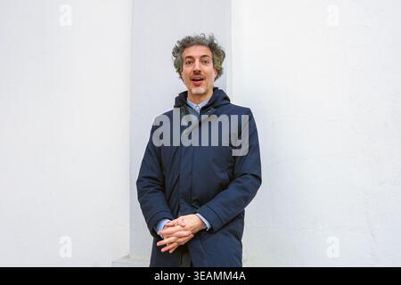 Homme aux cheveux bouclés debout contre un mur blanc, exprimant la surprise avec la bouche ouverte et les mains serrées Banque D'Images