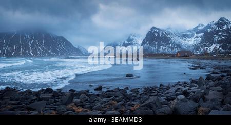 Les vagues s'écrasent doucement sur la rive rocheuse de Skagsanden Beach alors que les montagnes enneigées se dressent sous un ciel nuageux, créant une scène hivernale époustouflante, Lofo Banque D'Images