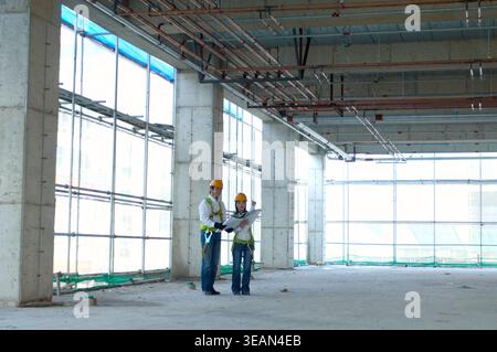 Deux ouvriers de la construction vêtus de casques de sécurité et de gilets de sécurité examinent les plans dans un grand entrepôt vide, au sol en béton et doté de hautes fenêtres. Banque D'Images
