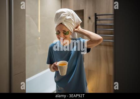 Rituel de beauté du matin de la femme heureuse d'âge moyen souriant quitte la salle de bain avec serviette sur la tête, tasse de thé Banque D'Images