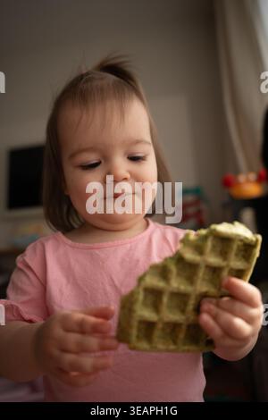 fille en bas âge curieuse dans une chemise rose à l'intérieur étudiant et tenant une collation gaufrée verte avec une expression concentrée et douce ; lumière naturelle douce, peu profonde Banque D'Images