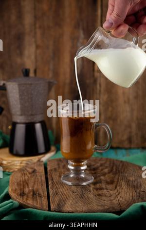 Main versant le lait frais de la carafe en verre dans la tasse à café avec Moka pot sur fond brun en bois rustique. Banque D'Images
