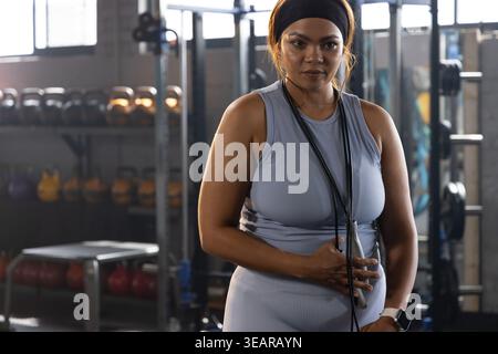 Femme afro-américaine debout dans un gymnase commercial portant des vêtements d'activité ajustés, corde à sauter, bandeau. Kettlebell, poids, banc, haltères, rack, lumière du jour Banque D'Images