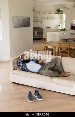 Homme incliné sur un canapé léger à la maison portant des écouteurs, une chemise à carreaux et un pantalon olive tenant le téléphone. Smartphone, sans fil, se prélasser, plan ouvert, maison, co Banque D'Images