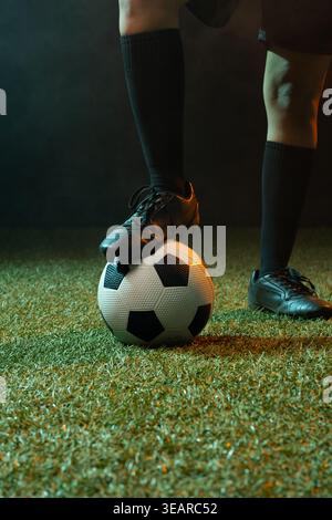 Femme afro-américaine équilibrant le crampon noir sur le ballon de football tout en portant des chaussettes hautes aux genoux sur le gazon. Athlète, chaussures, gros plan, dramatique, studio, brume, Banque D'Images