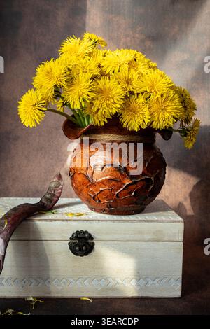 Nature morte verticale de pissenlits jaunes dans un pot en argile rustique reposant sur une boîte en bois blanc vintage. Composition florale élégante avec des tons chauds. Banque D'Images
